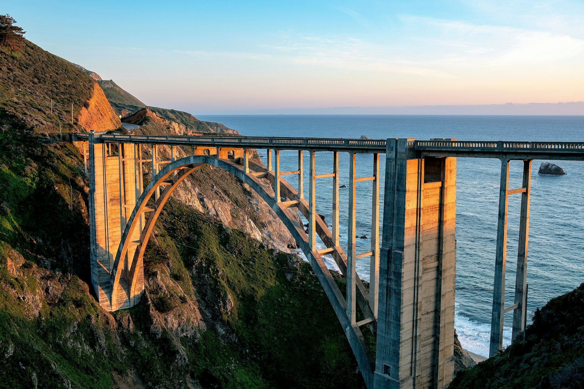 Arched concrete bridge spanning a coastal cliff with ocean views at sunset.
