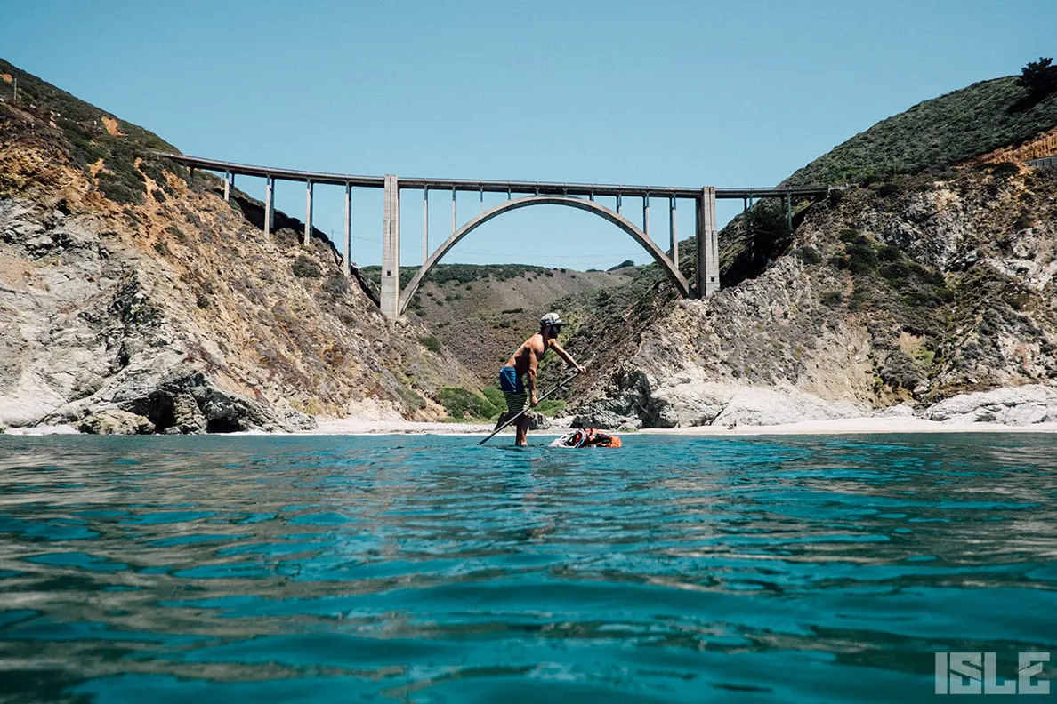A person paddleboarding on clear water beneath a bridge with rocky hills in the background.