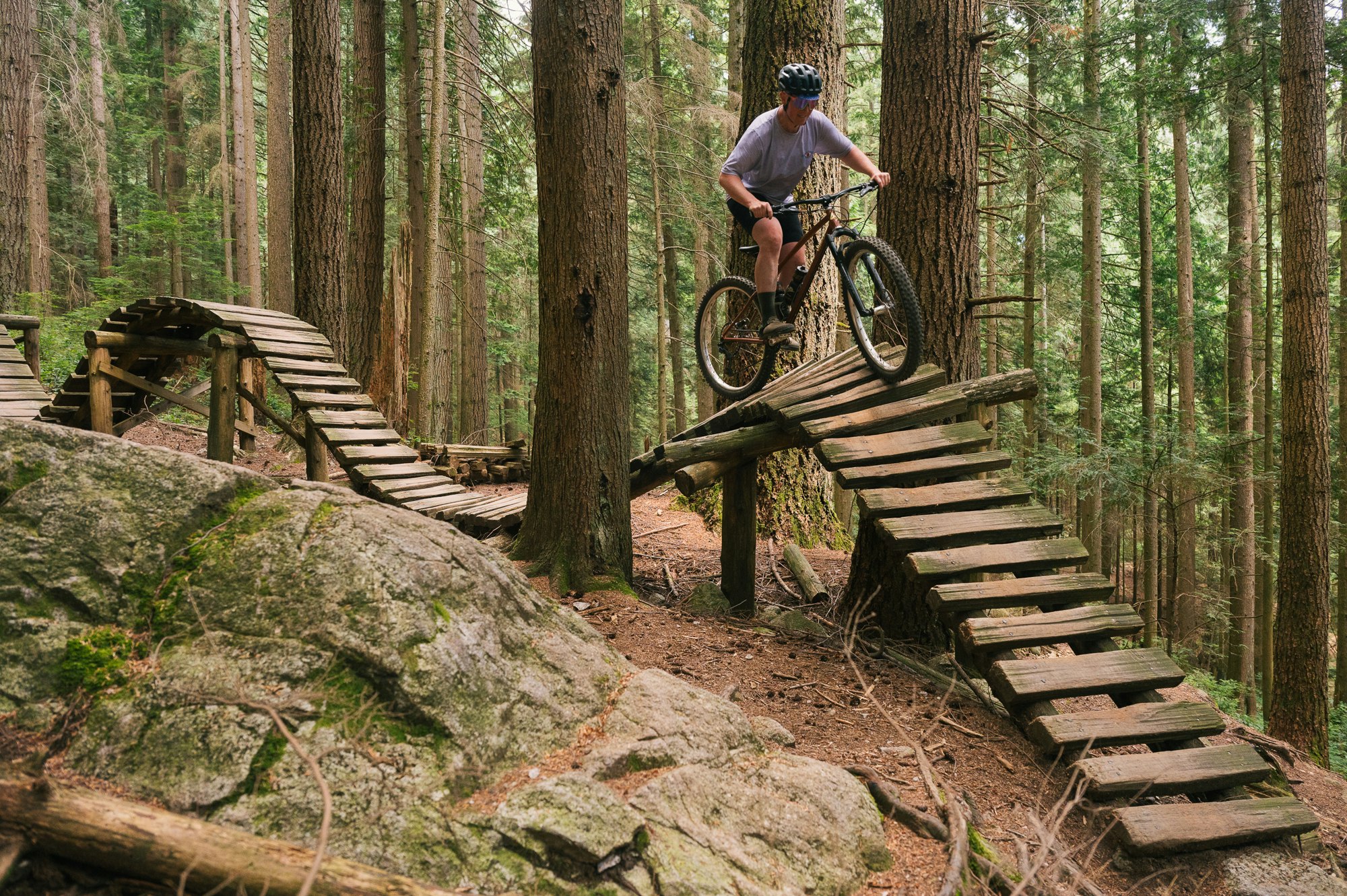 A mountain biker rides over wooden ramps in a forest, navigating a trail with trees and rocks around.