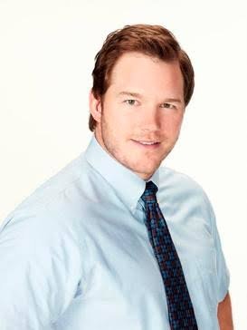 A man in a light blue shirt and patterned tie smiles against a plain background, showcasing a professional appearance.